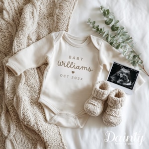 May include: A cream-coloured baby onesie with the text "Baby Williams" and a heart, along with "OCT 202X". Knitted baby booties and an ultrasound image are placed nearby. The items are arranged on a white surface with a beige knitted blanket.
