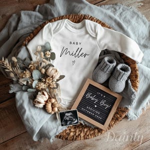 May include: A white baby bodysuit with the name "Miller" and a heart graphic, next to grey knitted baby booties and a letter board saying "It's a Baby Boy! Arriving October 202X". The display includes dried flowers and a sonogram image.