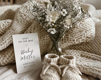 May include: A pregnancy announcement card with the text "Baby on the Way" and "Baby Miller arriving Oct 202X" is displayed next to a pair of cream-colored knitted baby booties. A bouquet of white flowers in a glass vase sits behind the card and booties.