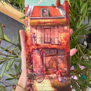 Puede incluir: Ilustración colorida de una librería con la palabra "LIBRUM" en el toldo. La imagen presenta un tejado de tejas rojas, una bicicleta y una persona leyendo en un balcón. La escena está llena de colores cálidos y exuberante follaje.