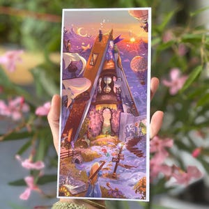 May include: A vibrant fantasy art print showing a house integrated into a large book. The artwork illustrates a sunset with hot air balloons, a crescent moon, and figures exploring the scene. The print is held in a hand.