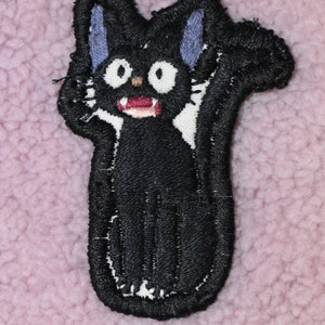 May include: Black embroidered patch of a cartoon cat with white fur and blue ears. The cat has a wide open mouth with white teeth and a red tongue.