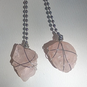 May include: Two rose quartz pendants. Each pendant features a rough-cut, light pink stone wrapped in silver-colored wire and suspended from a silver ball chain. The pendants are displayed against a white background.