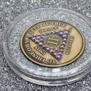 May include: A bronze coin with a triangle design featuring the words "To Thine Own Self Be True", "Service", "Unity", "Recovery". The triangle is decorated with iridescent rhinestones and the number "II" is in the center.