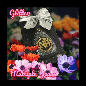 Peut inclure: Un porte-clés noir et argenté à paillettes avec un logo NA doré. Le porte-clés est entouré de fleurs roses, blanches et violettes. Le texte "Glitter Multiple Years" est écrit en lettres roses.