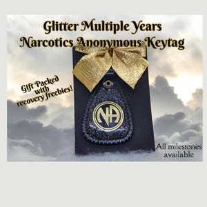 Pode incluir: Chaveiro de couro preto com acabamento de glitter dourado e um círculo dourado com as letras NA em preto. O chaveiro está amarrado com uma fita dourada e o texto "Glitter Multiple Years Narcotics Anonymous Keytag" está na parte superior da imagem. O texto "Gift Packed with recovery freebies!" está no canto inferior esquerdo da imagem. O texto "All milestones available" está no canto inferior direito da imagem.