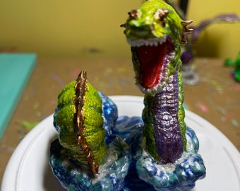 May include: A green and purple sea serpent figurine with gold accents, with its mouth open and red tongue showing, standing on a blue and white wave base.