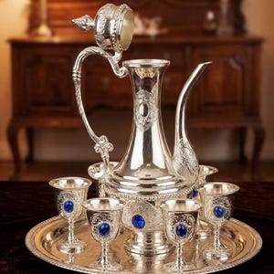 May include: A silver-colored tea set with ornate detailing, including a tall pitcher with a curved spout and handle, a tray, and six small goblets. Each goblet features a blue gemstone. The set is displayed on a dark surface.