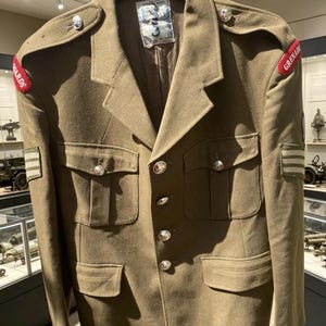 Peut inclure: Veste militaire vert olive avec des boutons argentés et des épaulettes. La veste comporte plusieurs poches et écussons, dont un avec l'inscription "GRENADE" en rouge. La veste est exposée dans un cadre muséal.