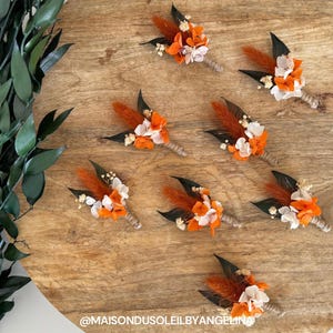 Op de afbeelding: Zeven oranje en witte bloemen boutonnières met bruine en zwarte veren op een houten oppervlak.