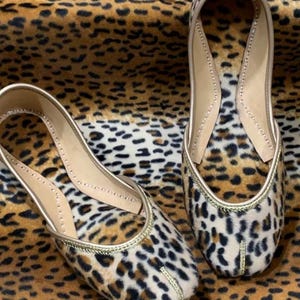 May include: A pair of flat shoes with a leopard print design and gold trim. The shoes are made of a soft, plush fabric and have a pointed toe.