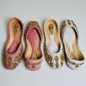 May include: Three pairs of flat shoes with gold embroidery and embellishments. The shoes are in pink, white, and cream colors.