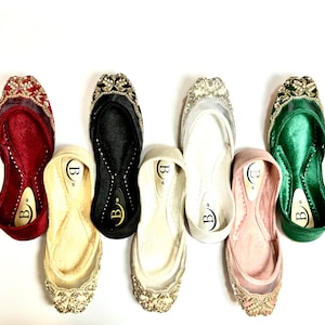 May include: Six pairs of women's flat shoes with jeweled embellishments. The shoes are in various colors, including red, black, white, pink, and green.