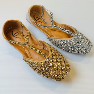 May include: Two pairs of gold and silver embellished flat shoes with a t-strap design. The shoes have a crescent moon logo with the letter "B" inside.