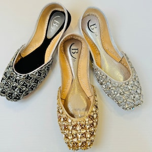 May include: Three pairs of embellished flat shoes in black, gold, and silver. Each shoe features a floral pattern of sparkling crystals. The interior of each shoe is a light color, and the brand logo "B5" is visible.