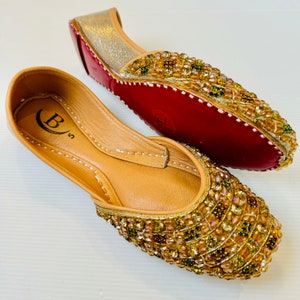 May include: A pair of gold embellished flat shoes with a red sole. The shoes have a pointed toe and are decorated with gold and iridescent beads.