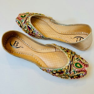 May include: A pair of gold embroidered flat shoes with colorful embellishments. The shoes have a pointed toe and a low heel. The inscription "B 5" is visible on the inside of the shoes.