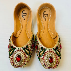 May include: A pair of white embroidered flat shoes with red and green embellishments. The shoes have a gold trim and are decorated with rhinestones. The shoes are size 5.