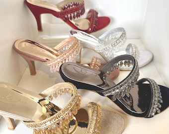 May include: A collection of five high-heeled mule sandals in red, silver, rose gold, black, and gold. Each shoe features a wide strap adorned with dangling crystal-like beads and a metallic insole stamped with the letter B and a size indicator.