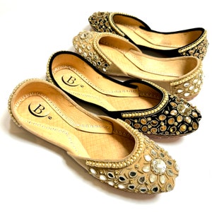 May include: Four pairs of embellished flats in a variety of colors. The shoes have a pointed toe and are decorated with mirrors and beads. The soles are tan and the uppers are black, gold, and tan. The shoes are arranged on a white surface.