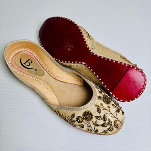 May include: A pair of gold embroidered flat shoes with red soles. The shoes have a floral design and are embellished with gold sequins.