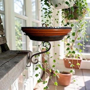 May include: A terracotta bird bath with a dark blue mosaic rim, mounted on a brown metal scrollwork bracket. The bracket features a clamp mechanism attached to a wooden surface, supporting the circular basin in a horizontal position.