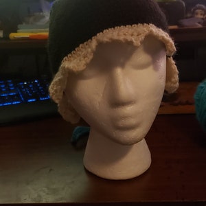 Może przedstawiać: Czarna czapka z białym haftowanym wykończeniem. Czapka jest na białej styropianowej głowie.