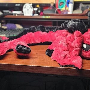 May include: A red and black plush dragon toy with a long, segmented body and a small head with black eyes. The dragon is lying on a wooden surface.