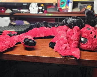 May include: A red and black plush dragon toy with a long, segmented body and a small head with black eyes. The dragon is lying on a wooden surface.