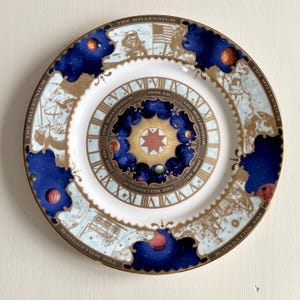 May include: A decorative plate with a white centre and a blue and gold rim. The plate features a celestial design with planets, stars, and the text "THE MILLENNIUM" and "2000 AD". The rim has a gold border.