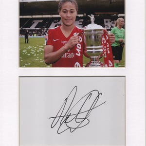 May include: Framed photograph of a woman in a red jersey holding a silver trophy with a red ribbon. The jersey has the word "arat" visible. Below is a white background with a black signature.