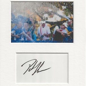 May include: A black and white signed photograph of a group of men celebrating with champagne. The men are wearing white shirts and hats. The photo is signed "Rickie Fowler".