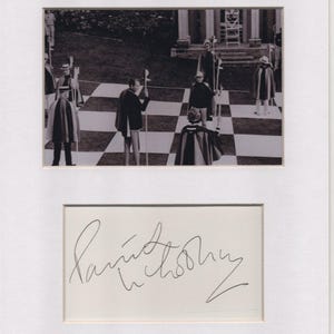 May include: Black and white photograph of a chess game in progress, with people dressed in capes and hats standing on a giant chessboard. Below the photo is a white card with a handwritten signature.