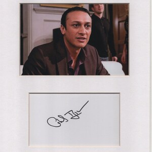 May include: A framed photograph featuring a man in a brown blazer and a purple shirt. Below the photo is a white rectangle with a black signature.