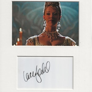 May include: A black and white signed photograph of a woman wearing a white dress and a tiara. The woman is looking directly at the camera. The background is a dark blue.