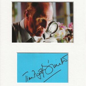 May include: A black and white signed photograph of Tim Roth, an actor, wearing a white shirt and tie. He is looking through a magnifying glass at a flower. The signature is on a blue background.