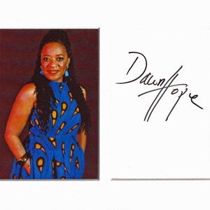 May include: A framed photograph featuring a woman in a blue dress with yellow patterns. The dress has a high neckline and sleeveless design. The woman is wearing earrings and a bracelet. Next to the photo is a white card with the handwritten signature "Dawn/foye".