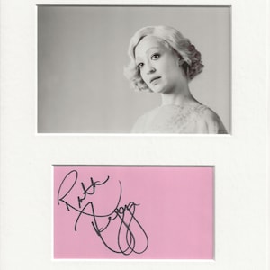 May include: Black and white photo of a woman with short, curly hair. She is wearing a light-colored dress and looking to the side. The photo is signed "Ruth Negga" in black ink on a pink background.