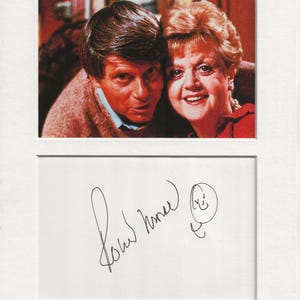 May include: A black and white photograph of two people, a man and a woman, smiling. The woman is wearing a red jumper and the man is wearing a brown jumper. The photo is signed "John Inman" with a smiley face.