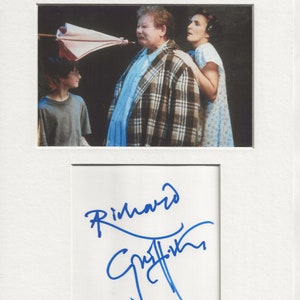 May include: A black and white photograph of a man and woman in a scene from a play. The man is wearing a plaid shirt and the woman is wearing a white dress. The man is holding an umbrella over his head. The photograph is signed "Richard Griffiths".