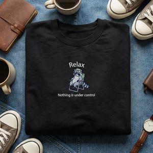 May include: Black sweatshirt with the word "Relax" above a cartoon raccoon wearing a hat and sunglasses, sitting in a chair. The phrase "Nothing is under control" is below the raccoon illustration.
