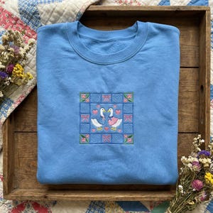May include: A pale blue sweatshirt with an embroidered square design featuring two geese. The geese are in a loving embrace, surrounded by pink hearts, bows, and floral accents. The sweatshirt is displayed on a wooden tray.