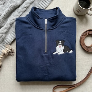 May include: Navy blue quarter-zip sweatshirt with a white and black Border Collie dog embroidery. The sweatshirt has a silver zipper and a high collar. A dog leash and a mug with a paw print are also visible.