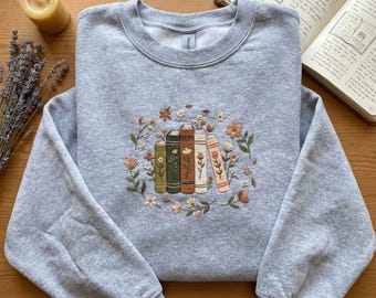May include: A light gray sweatshirt with an embroidered design of stacked books surrounded by flowers. The books are in shades of green, brown, and cream. A candle, dried lavender, and an open book are in the background.