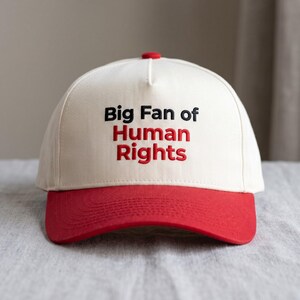 May include: A cream-coloured baseball cap with a red brim and a red button on top. The front features embroidered text that reads Big Fan of in black, with the words Human Rights embroidered in red underneath the black text.