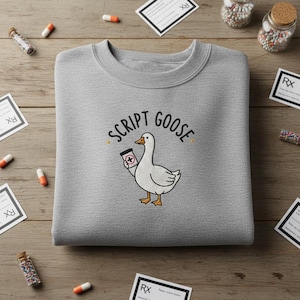 May include: A heather gray sweatshirt featuring the phrase "SCRIPT GOOSE" above an illustrated goose holding a prescription bottle. The goose is white with yellow feet and a yellow beak. The design is surrounded by prescription-related items.