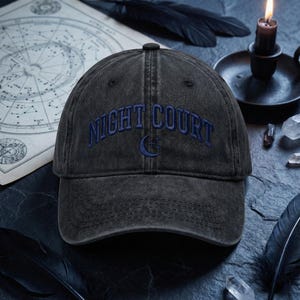 May include: A dark gray baseball cap with the embroidered text "NIGHT COURT" in blue. A crescent moon and cross design is embroidered below the text. The hat has a curved brim and a fabric strap closure.