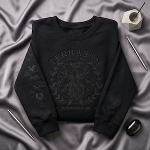 May include: Black crew neck sweatshirt with the word "TERRASEN" above a circular design featuring a crown, mountains, and a floral wreath. The phrase "TO WHATEVER END" is below the design. The sleeves have embroidered details.