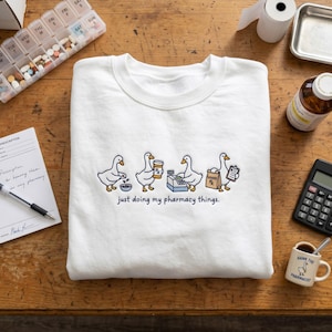 May include: A white crewneck sweatshirt featuring a graphic of four geese performing pharmacy tasks, including counting pills, holding a prescription bottle, using a cash register, and carrying a bag labeled Goose Rx, with the text just doing my pharmacy things.