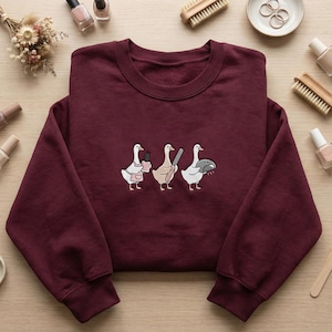 May include: A maroon sweatshirt with an embroidered design of three geese. Each goose is shown with a different item: nail polish, a knife, and a pie. The sweatshirt has a crew neck and long sleeves.